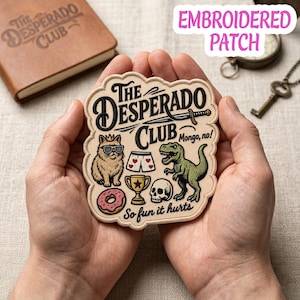 May include: Embroidered patch with the text "The Desperado Club" and various cartoon illustrations, including a cat, dinosaur, and skull. The patch is held in a person's hands.