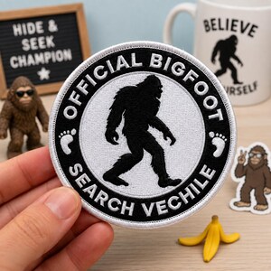 May include: A circular embroidered patch featuring a black silhouette of Bigfoot in the center on a white background. The black border contains the white text OFFICIAL BIGFOOT SEARCH VECHILE with two small white footprints on the sides.