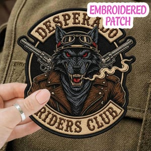 May include: Embroidered patch featuring a wolf wearing goggles and a brown leather jacket, with the words "DESPERADO RIDERS CLUB" and two revolvers. The wolf has red eyes and is smoking. The patch is black, brown, and tan.