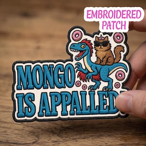 May include: Embroidered patch with the text "MONGO IS APPALLED". The patch features a blue dinosaur and a brown cat wearing sunglasses. The patch is surrounded by pink donuts.