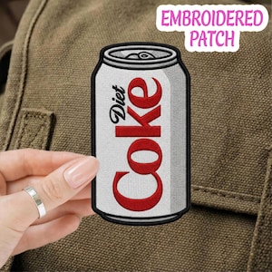 Peut inclure: Un écusson brodé en forme de canette de soda argentée avec une bordure noire. Le texte Diet Coke est cousu en rouge sur le devant de la canette. L'écusson est placé sur une surface en tissu marron texturé, avec un détail de languette sur le dessus.