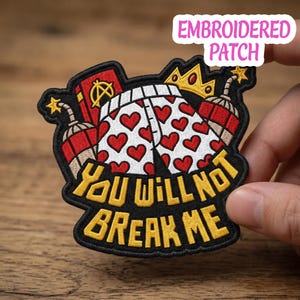 Peut inclure: Écusson brodé avec une bordure noire, représentant un motif de fesses couvertes de cœurs, une couronne et de la dynamite. Le texte "YOU WILL NOT BREAK ME" est en jaune. L'écusson est tenu par une main.