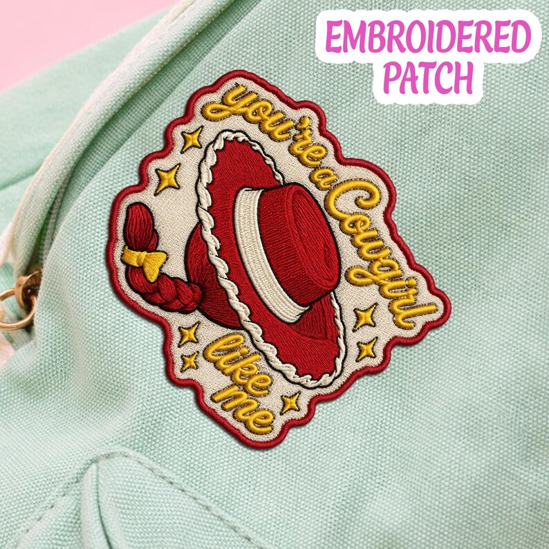 May include: An embroidered patch featuring a red cowboy hat with a white band and a red braided hair detail. The text "you're a cowgirl like me" is stitched in yellow cursive script around the hat, accented by small yellow stars on a cream background.