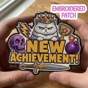 Puede incluir: Parche bordado con un gato gruñón con corona, el texto "NEW ACHIEVEMENT!" y gráficos temáticos de juegos. El parche incluye una calavera, un trofeo, una bomba, un rayo y una espada. Los colores son morado, dorado y gris.