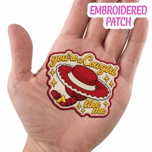 May include: An embroidered patch featuring a red cowgirl hat with a white band and a red braided ponytail with a yellow bow. The text reads you're a cowgirl like me in yellow cursive script, surrounded by small yellow stars on a cream background.