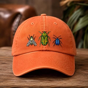 May include: An orange baseball cap featuring embroidered insect designs on the front panel. The embroidery includes a white and black fly, a green beetle with intricate patterns, and a small solid blue beetle, all centered above the curved brim.