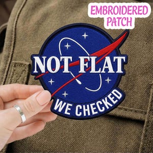 May include: A circular embroidered patch featuring a dark blue background with white stars and a white orbital line. A red swoosh crosses the center. The text reads NOT FLAT in white letters across the middle and WE CHECKED in white at the bottom.