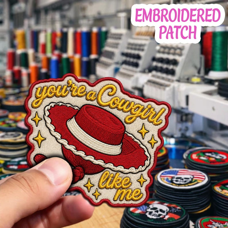 May include: An embroidered patch featuring a red cowboy hat with a white band, surrounded by yellow star icons. The text reads you're a Cowgirl like me in yellow cursive script against a cream background with a red border and scalloped edges.