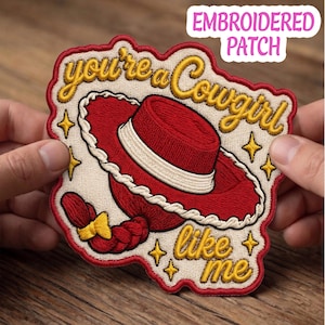 May include: An embroidered patch featuring a red cowboy hat with a white band and a red braided hair detail with a yellow bow. The text reads you're a Cowgirl like me in gold cursive script, surrounded by small gold stars on a cream background.