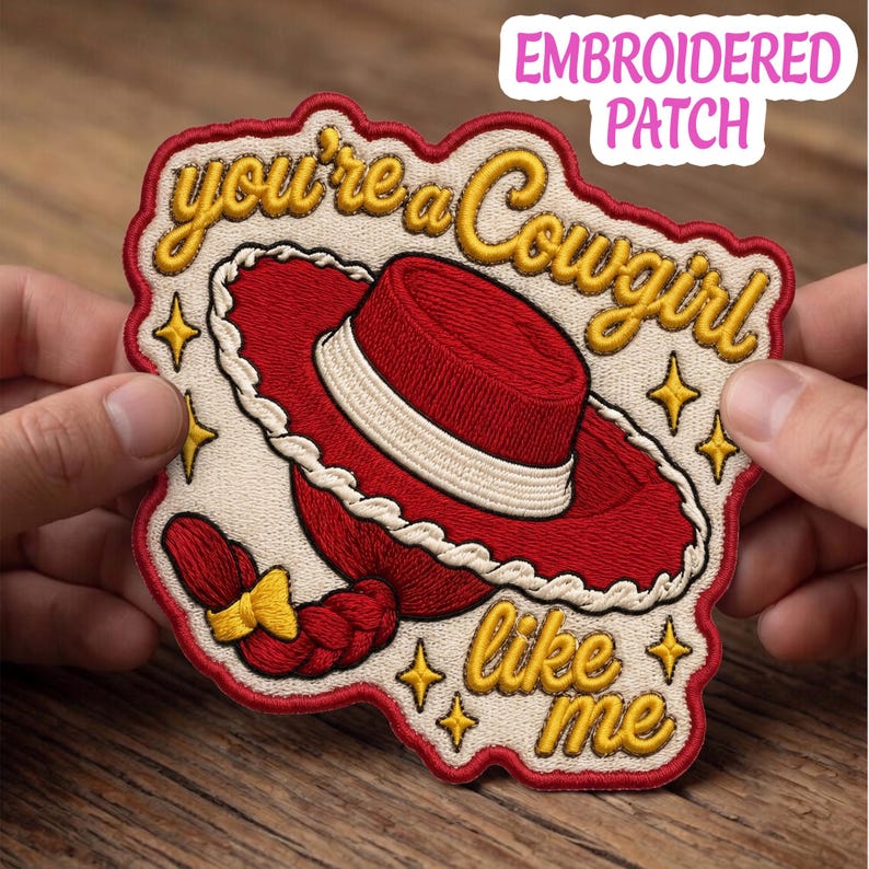 May include: An embroidered patch featuring a red cowboy hat with a white band and a red braided hair detail with a yellow bow. The text reads you're a Cowgirl like me in gold cursive script, surrounded by small gold stars on a cream background.