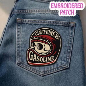 May include: An embroidered patch featuring a cream-colored skull wearing a red and black motorcycle helmet. The patch has black borders and includes the text CAFFEINE & GASOLINE in cream-colored lettering. It is attached to a blue denim pocket.