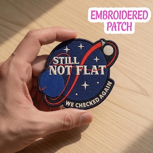 May include: Embroidered patch with a space-themed design. The patch features a blue planet, a red curved line, and the text "STILL NOT FLAT" and "WE CHECKED AGAIN" in white. The patch is held in a hand.