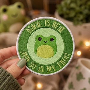 May include: A circular embroidered patch featuring a light green frog with dark eyes and a pale yellow belly at the center. The patch has a dark green border with the text "MAGIC IS REAL AND SO IS MY FROG!" stitched in light green thread.