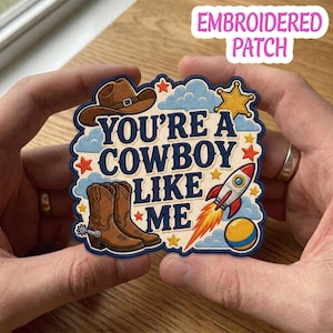 May include: An embroidered patch featuring the text "YOU'RE A COWBOY LIKE ME" in dark blue lettering. The design includes a brown cowboy hat, brown cowboy boots, a red and white rocket with orange flames, a yellow star, and blue clouds.