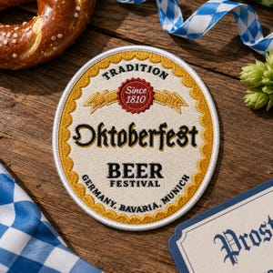 May include: A round embroidered patch with a cream-colored center and a scalloped gold border. The text reads Oktoberfest, Beer Festival, Tradition, Since 1810, Germany, Bavaria, Munich. The design features a red circular emblem and gold ribbons.