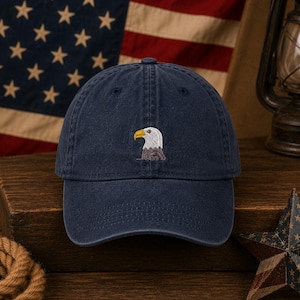 May include: A navy blue baseball cap featuring a small, centered embroidery of a bald eagle head in profile. The eagle has a white head, a yellow beak, and brown neck feathers. The cap has a structured crown and a curved brim, with visible stitching.