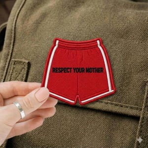May include: A red patch in the shape of shorts with white trim and the words "RESPECT YOUR MOTHER" in black. The patch is being held up against a green background.