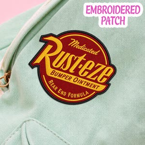 May include: A circular embroidered patch featuring a red background with a yellow border and yellow text that reads Rust-eze Medicated Bumper Ointment Rear End Formula. The patch is attached to a light mint green fabric surface.