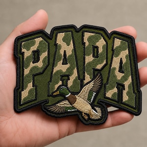 May include: Embroidered patch with the word "PAPA" in green and camouflage lettering. A detailed illustration of a flying duck is featured below the text, with brown, green, and white feathers. The patch has a black border.