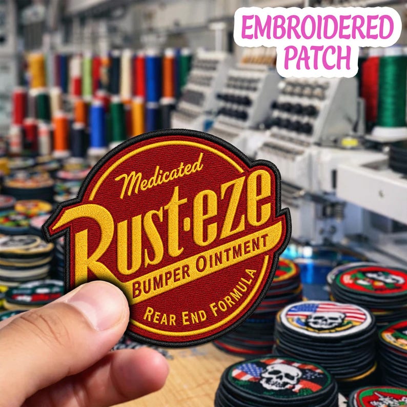 May include: A circular embroidered patch featuring a red background with a yellow border and yellow text that reads Medicated Rust-eze Bumper Ointment Rear End Formula. The patch has a black stitched edge and is held against a blurred background.