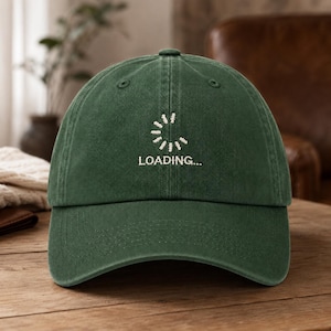 May include: A dark forest green baseball cap featuring an embroidered white circular loading icon and the text "LOADING..." centered on the front panel. The cap has a structured crown, a curved brim, and stitched eyelets for ventilation.