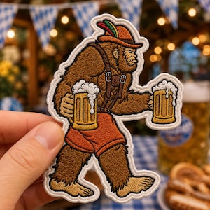 May include: An embroidered patch featuring a brown Bigfoot character wearing a brown hat with a red and green feather, brown suspenders, and orange shorts. The character holds two yellow beer mugs with white foam in each hand.