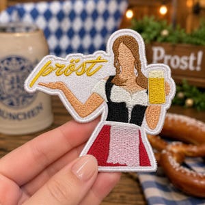 May include: An embroidered patch featuring a person with brown hair wearing a white blouse, black bodice, and red skirt, holding a yellow beer mug. The text "pröst" is stitched in yellow cursive script above the person's outstretched arm.