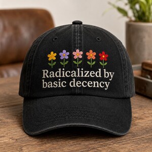 May include: A black baseball cap featuring an embroidered row of five colorful flowers in yellow, purple, pink, orange, and red. Below the flowers, the text "Radicalized by basic decency" is embroidered in white thread across the front panel.