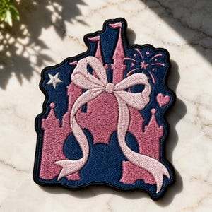 May include: Embroidered patch featuring a pink castle with a large pink bow and flowing ribbons, set against a navy blue background. A star, heart, and fireworks are also included in the design. The patch has a black border.