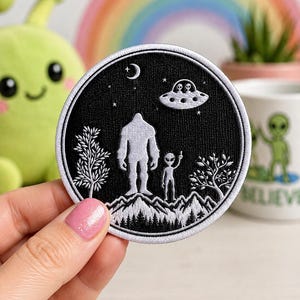 May include: A circular embroidered patch featuring a black background with a white silhouette of Bigfoot standing next to an alien on a mountain range. Above them, a flying saucer with two aliens inside floats under a crescent moon and stars.