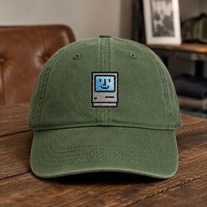 May include: An olive green baseball cap featuring an embroidered pixel art graphic of a vintage computer with a light blue screen and a smiling face. The cap has a structured crown, a curved brim, and stitched eyelets for ventilation.
