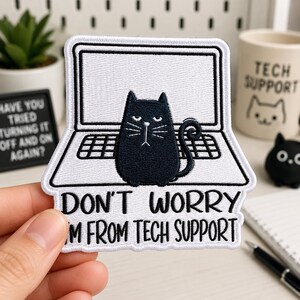 May include: A white and black embroidered patch featuring a black cat sitting in front of an open laptop. The text below the laptop reads, "DON'T WORRY I'M FROM TECH SUPPORT." The patch has a white border and is shaped to follow the outline of the design.