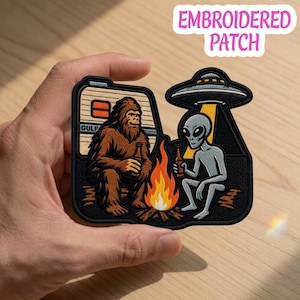 May include: An embroidered patch featuring a brown Bigfoot and a grey alien sitting by a campfire with a bottle in each hand. Above the alien is a UFO with a yellow beam of light. In the background is a camper trailer. The patch has a black border.
