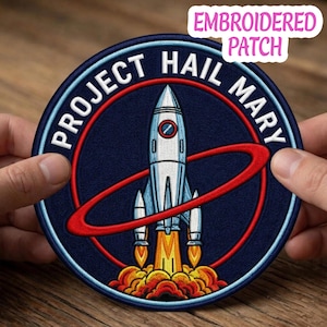 May include: A circular embroidered patch featuring a central silver rocket ship with orange and yellow flames at the base. A red ring encircles the rocket against a dark blue background. The text PROJECT HAIL MARY is stitched in white capital letters.