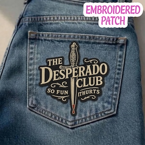 Könnte beinhalten: Gestickter Aufnäher auf Jeansstoff. Der Aufnäher zeigt die Worte "THE DESPERADO CLUB" mit einer Dolch-Illustration. Der Text "SO FUN IT HURTS" ist ebenfalls enthalten. Der Aufnäher ist schwarz, beige und rot.