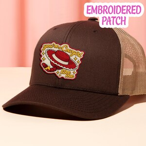 May include: A dark brown trucker hat with a tan mesh back and an embroidered patch on the front. The patch features a red cowboy hat, a yellow braid, and the text "you're a cowgirl like me" surrounded by small yellow star icons.