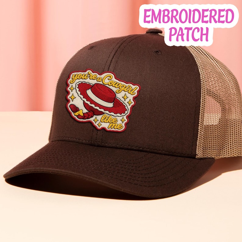 May include: A dark brown trucker hat with a tan mesh back and an embroidered patch on the front. The patch features a red cowboy hat, a yellow braid, and the text "you're a cowgirl like me" surrounded by small yellow star icons.
