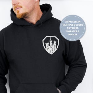 May include: Black hoodie with a front pocket and a white chess piece emblem on the chest. The emblem features a king, queen, and other chess pieces inside a shield shape. The hoodie is available in multiple colors as a t-shirt, sweater, and hoodie.