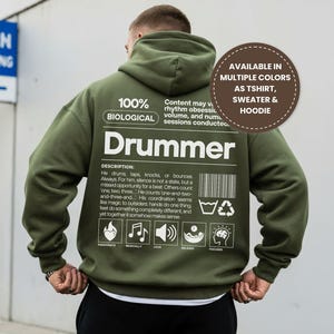 May include: An olive green hoodie featuring the word "Drummer" printed on the back, accompanied by text and graphics. The hoodie includes a hood and a white drawstring. The text on the back says "Available in multiple colours as T-shirt, sweater & hoodie."