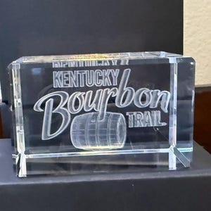 Puede incluir: Un bloque de vidrio rectangular transparente con las palabras "Kentucky Bourbon Trail" grabadas en la parte delantera. El diseño incluye un gráfico de barril. El bloque se muestra sobre una superficie negra.