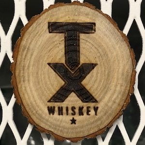 Pode incluir: Uma base de copo redonda de madeira com um design queimado. O design inclui as letras "TX" e a palavra "WHISKEY" com uma estrela abaixo. A base de copo tem um grão de madeira natural e uma borda marrom escura.