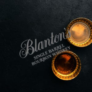 Pode incluir: Dois copos de whisky com bordas douradas cheios de líquido âmbar estão sobre uma superfície preta texturizada. Os copos estão posicionados perto do texto "Blanton's Single Barrel Bourbon Whiskey" em uma fonte estilizada.