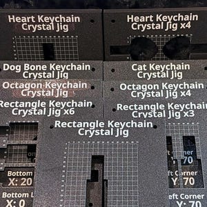 Puede incluir: Una colección de plantillas rectangulares de cristal para llaveros en gris oscuro con texto impreso en blanco y patrones de cuadrícula. Las plantillas están etiquetadas para varias formas, incluyendo corazón, hueso, gato, octágono y rectángulo.