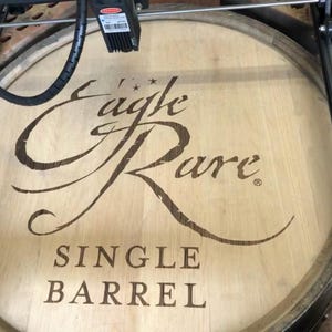 Pode incluir: Tampa de barril de madeira clara com as palavras "Eagle Rare Single Barrel" gravadas na madeira. O texto está em uma fonte estilizada. A tampa parece fazer parte de um barril maior.