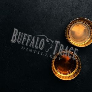 Op de afbeelding: Twee whiskyglazen met gouden rand, gevuld met amberkleurige vloeistof, staan op een zwarte ondergrond. Het logo van Buffalo Trace Distillery is subtiel zichtbaar op de achtergrond. De glazen zijn gevuld met een donker amberkleurige vloeistof.