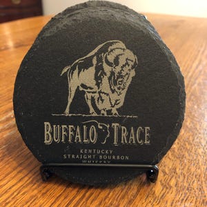 May include: A set of black slate coasters with a silver-colored bison graphic and the words "BUFFALO TRACE" and "KENTUCKY STRAIGHT BOURBON WHISKEY" etched on them. The coasters are displayed in a black metal holder.