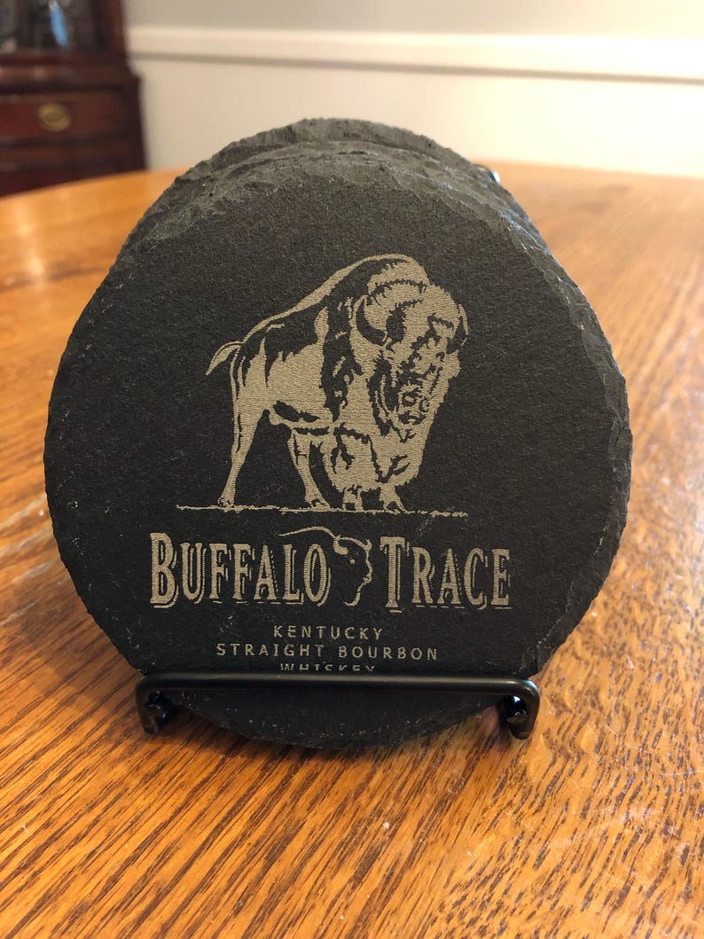 May include: A set of black slate coasters with a silver-colored bison graphic and the words "BUFFALO TRACE" and "KENTUCKY STRAIGHT BOURBON WHISKEY" etched on them. The coasters are displayed in a black metal holder.