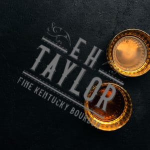 Pode incluir: Dois copos de whisky com bordas douradas cheios de líquido âmbar estão em uma superfície escura. Os copos estão posicionados perto do texto "EH TAYLOR FINE KENTUCKY BOURBON" em uma fonte estilizada.