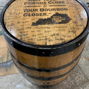 Pode incluir: Um barril de bourbon de madeira com faixas de metal preto. A parte superior do barril apresenta o texto "KEEP YOUR FRIENDS CLOSE & YOUR BOURBON CLOSER." e o logotipo Kentucky Bourbon Trail. O barril está assinado.