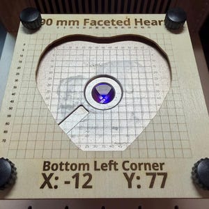 Könnte beinhalten: Eine helle Holzschablone mit herzförmigem Ausschnitt, einem Gittermuster und einer zentralen kreisförmigen Linse. Der Text lautet 90 mm Faceted Heart, Bottom Left Corner, X: -12, Y: 77. Vier schwarze Knöpfe befinden sich an den Ecken.